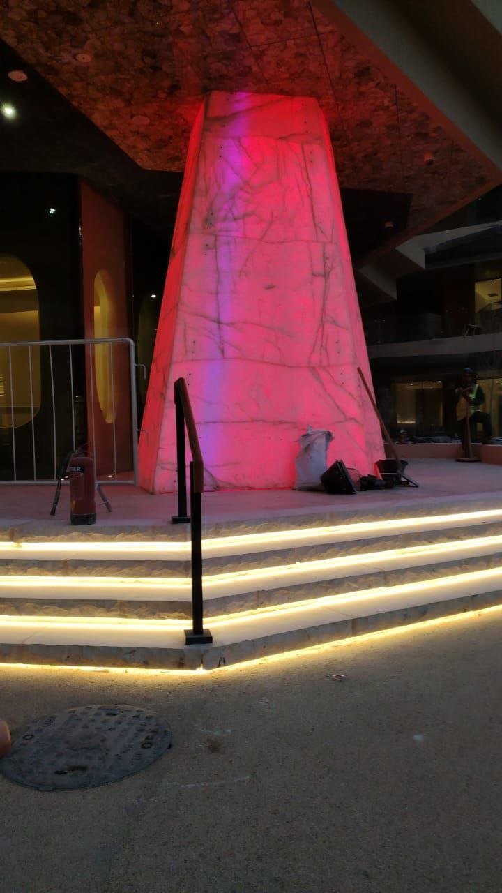 A tall marble obelisk lit in deep pink and red from within, with warm lighting wrapping the stair base.